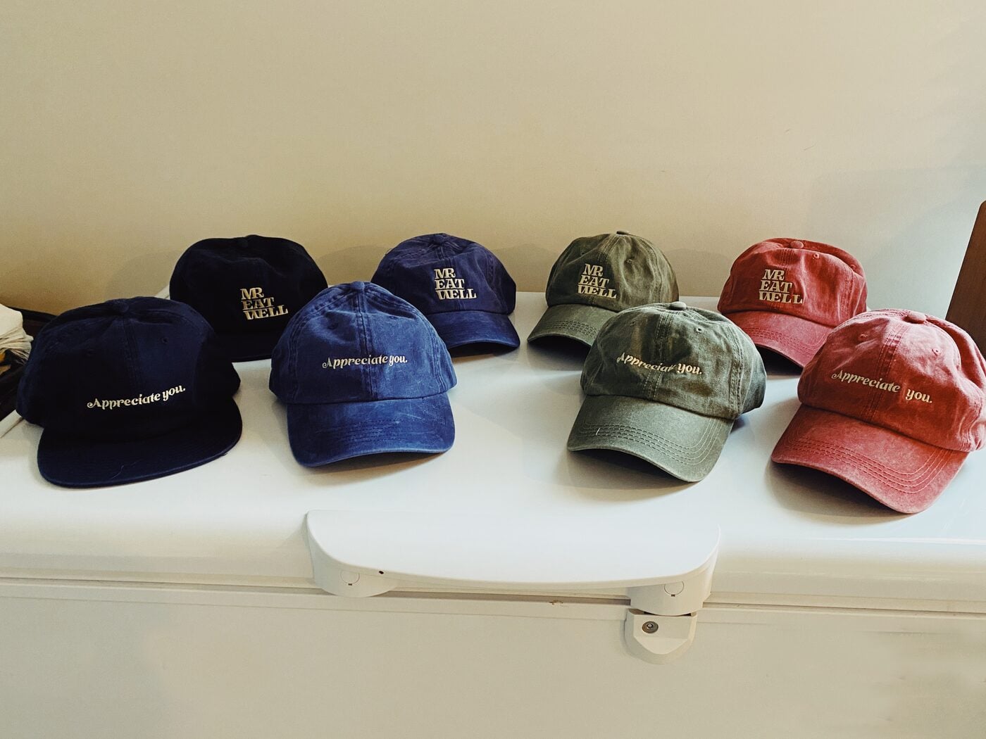 Seven washed dad caps laid on a white chest freezer, four embroidered “Appreciate you.”