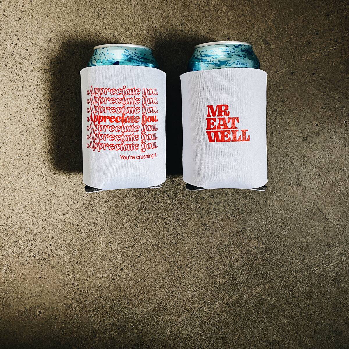 Two white koozies on concrete, one printed with a repeating “Appreciate you.” stack in red