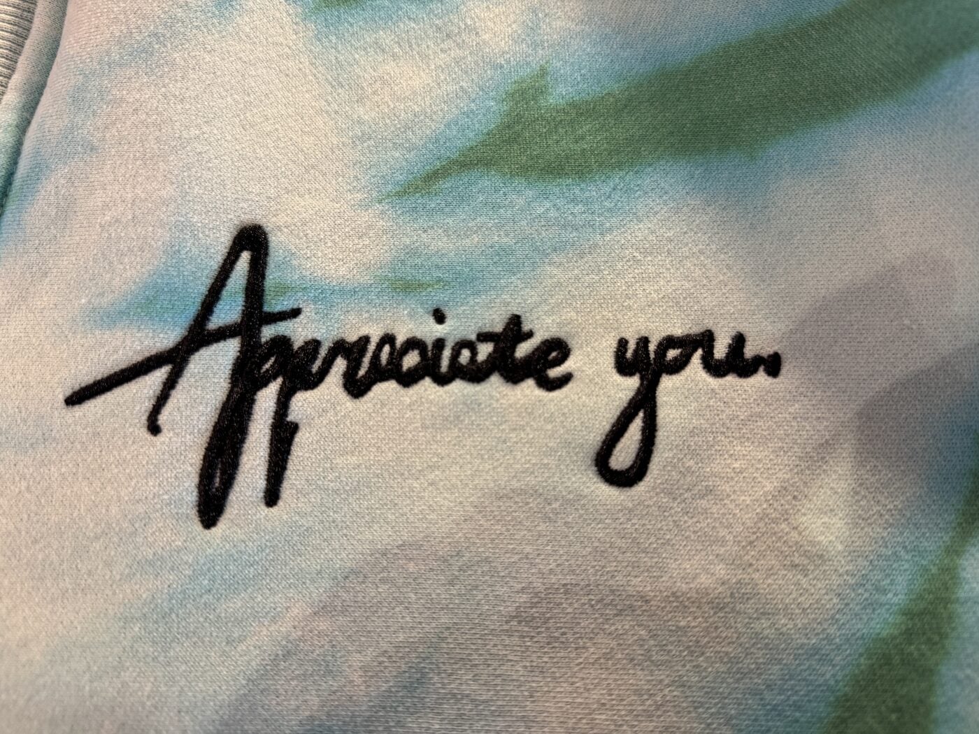 Macro of “Appreciate you.” chain-stitch embroidered in black thread on pale tie-dye fleece