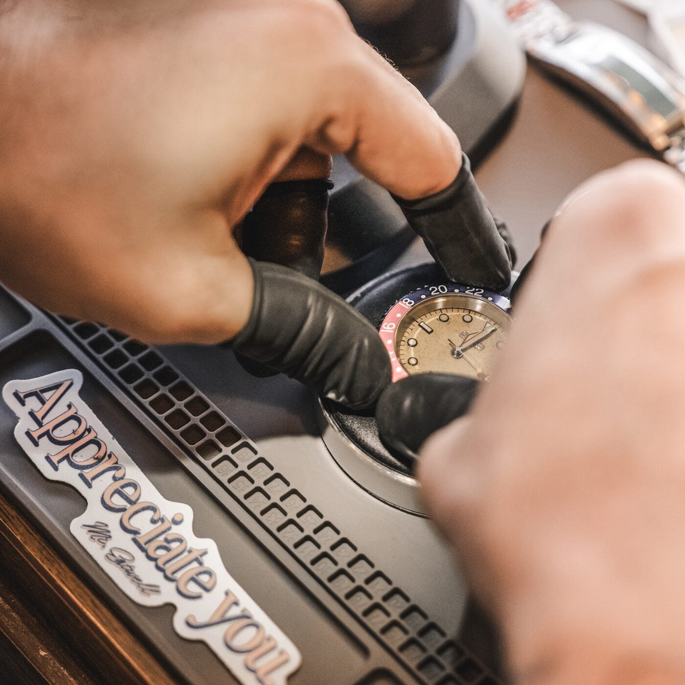 Gloved hands working on a motorcycle with a GMT watch on the wrist and a die-cut “Appreciate you.” sticker on the bodywork