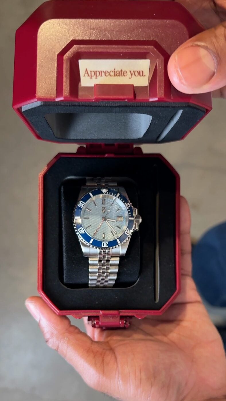 Hands holding an open burgundy watch case with the “Appreciate you.” logo on the lid and a blue-dial diver seated below