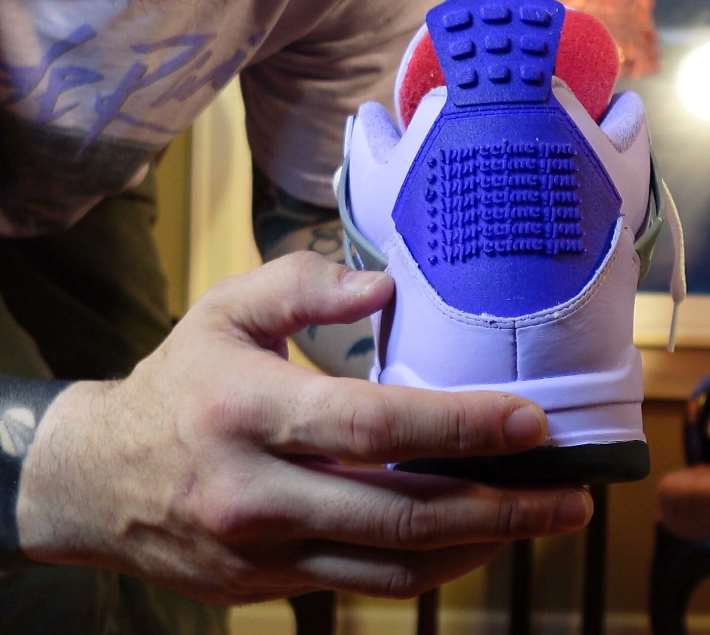 A hand holding a hand-painted purple Jordan 4, heel toward camera, with “appreciate you.” debossed in repeating rows across the blue nubuck heel panel